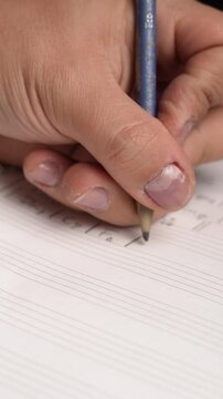 Close-up of a hand holding a blue pencil, writing on a lined sheet of paper. The structured lines suggest it might be music notation or a formatted document used for composition.