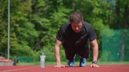 Athletic man doing push-ups on red running track with water bottle nearby. Outdoor workout session. Person training on stadium. Active lifestyle, sport and fitness concept