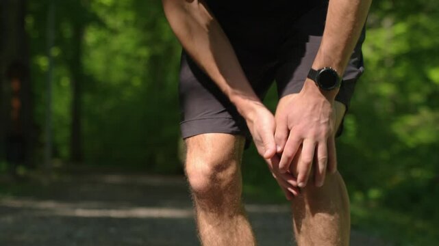 Runner holding knee in pain during workout in summer park. Man has injury while running. Male athlete suffering from knee pain after sport exercises