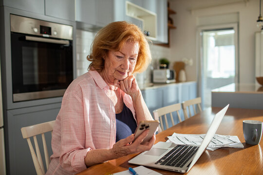 Concerned senior woman looking at smartphone while managing bills at home with laptop