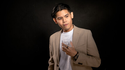 Asian man in beige blazer and white t-shirt holding a transparent glass, looking away with contemplative expression in studio with dark background, perfect for lifestyle or branding visuals