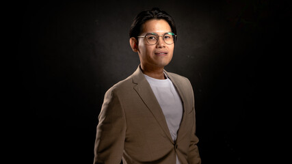 Fototapeta premium Asian man wearing glasses, beige blazer, and white shirt standing in studio with dark background, looking forward with confident posture, perfect for business, branding, or marketing visuals