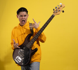 Obraz premium Asian man in a yellow hoodie playing an electric bass guitar against a bright yellow background. Captured mid-performance with a focused expression and casual modern style