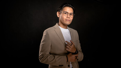 Asian man wearing beige blazer and white shirt standing in studio with dark background, confidently pointing to chest, ideal for branding, leadership, or creative business expression