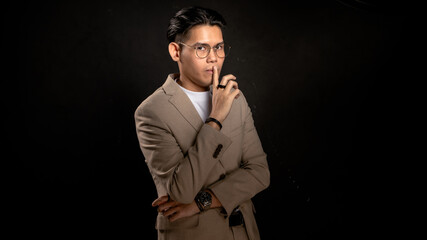 Asian man in beige blazer and glasses posing with finger on lips in studio against dark background, making quiet gesture, suitable for secrecy, confidentiality, or creative concept visuals