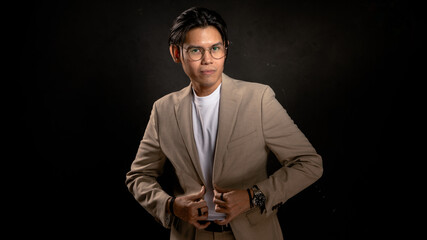 Asian man in beige blazer and white t-shirt adjusting jacket while looking sideways in studio with dark background, expressing confidence and style, ideal for business, fashion, or branding use