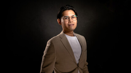 Obraz premium Asian man wearing glasses, beige blazer, and white shirt standing in studio with dark background, looking forward with confident posture, perfect for business, branding, or marketing visuals