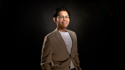 Obraz premium Asian man wearing glasses, beige blazer, and white shirt standing in studio with dark background, looking forward with confident posture, perfect for business, branding, or marketing visuals
