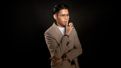 Asian man in beige blazer and glasses posing with finger on lips in studio against dark background, making quiet gesture, suitable for secrecy, confidentiality, or creative concept visuals