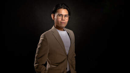 Asian man wearing glasses, beige blazer, and white shirt standing in studio with dark background, looking forward with confident posture, perfect for business, branding, or marketing visuals