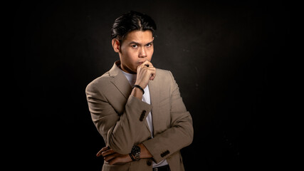 Asian man in beige blazer and white shirt posing with hand on mouth in thoughtful expression, standing against dark background, ideal for concepts like thinking, strategy, or decision-making