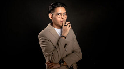Asian man in beige blazer and glasses posing with finger on lips in studio against dark background, making quiet gesture, suitable for secrecy, confidentiality, or creative concept visuals