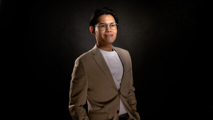 Asian man wearing glasses, beige blazer, and white shirt standing in studio with dark background, looking forward with confident posture, perfect for business, branding, or marketing visuals