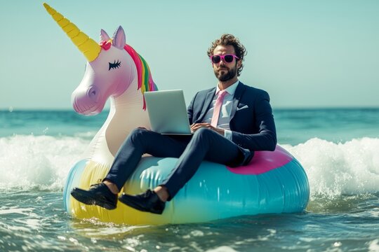 Businessman wearing suit and sunglasses using laptop while floating on inflatable unicorn in ocean, enjoying work-life balance