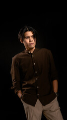 Fototapeta premium Asian man in a brown shirt and light trousers standing with hands in pockets, gazing sideways in a moody studio portrait with black background, expressing calm thought and modern masculinity