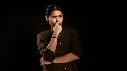 Asian man in dark brown shirt standing in studio with dark background, touching lips with thoughtful expression, perfect for concept visuals related to thinking, planning, or personal reflection