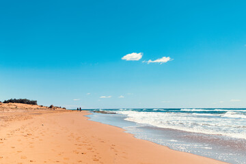 Wide view of Calblanque beach in Murcia, Spain with smooth orange sand, foamy shoreline, clear sky, and distant walkers creating a sense of vastness and peaceful isolation