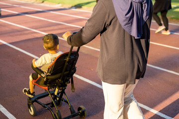 Obraz premium Back view of a woman in hijab pushing a stroller with a child on an athletic track, morning sunshine highlighting an active, healthy, and family-focused outdoor lifestyle.