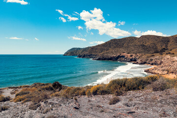Obraz premium Tranquil bay in Calblanque, Murcia, Spain with golden beach, lush coastal vegetation, turquoise sea, and dramatic hills under a bright sunny sky with scattered clouds