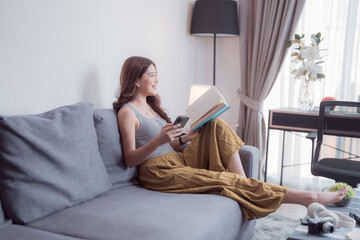 Young woman is sitting comfortably on a gray sofa in her modern living room, engrossed in a book...