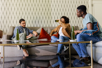 Relaxing male friends sitting on sofa at home, holding green beer bottles on glass coffee table