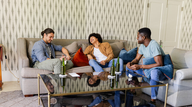 Laughing diverse male friends relaxing on sofa in living room, with green glass beer bottles