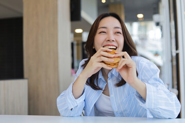 A cheerful young Asian woman is smiling brightly while holding a burger with both hands, ready to take a bite. She is enjoying her meal at a modern cafe or restaurant.