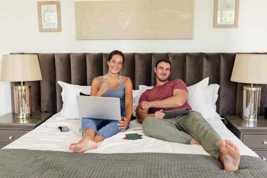 Relaxing couple reclining on king-size bed in bedroom, holding laptop and tablet with passports