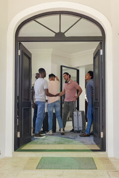 Greeting diverse male friends hugging and shaking hands at modern home entrance, with suitcases