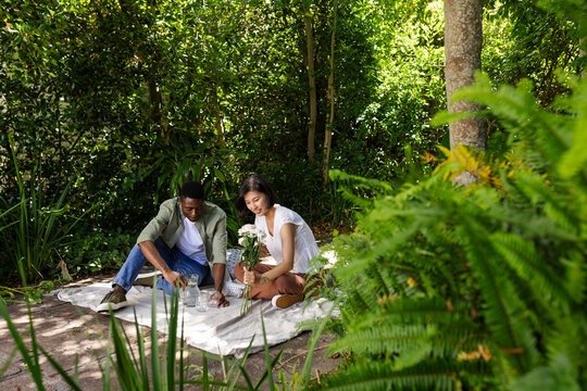 Diverse couple sharing picnic in garden with white picnic blanket, pitchers and flower bouquet