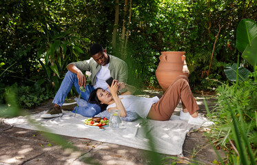 Relaxing Asian and African American couple lying on garden blanket, holding smartphone and rose