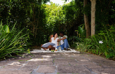 Smiling diverse couple sitting on blanket on stone path in garden, with pillows, pitcher, snacks
