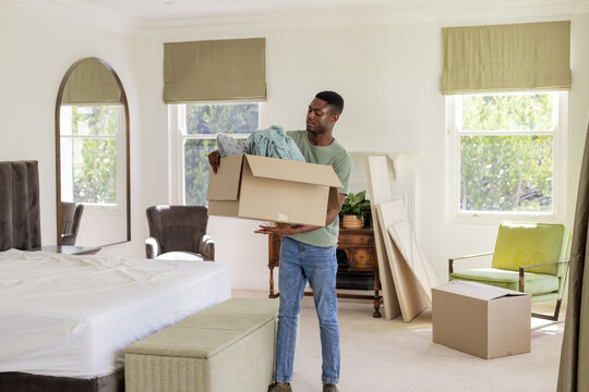Carrying African American man holding cardboard box of clothes in bedroom, with mirror and ottoman