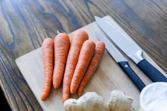 Light cutting board is resting on kitchen table, displaying carrots, knives and ginger root