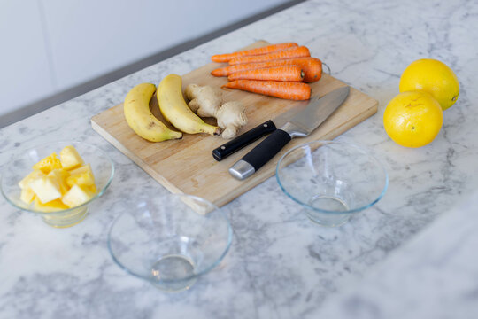 Wooden cutting board featuring ginger, carrots on marble countertop with pineapple bowl and knives - Powered by Adobe