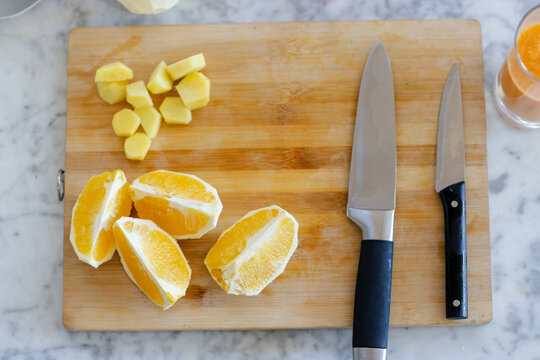 Wooden cutting board is holding orange wedges and diced ginger on marble counter near knives, glass