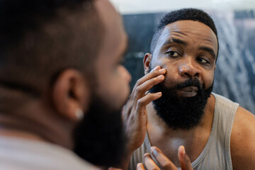 African American man examining skin, touching cheek in bathroom mirror, with wet glass shower door