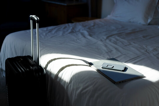 Black suitcase standing in bedroom, casting handle shadow on bed with laptop and smartphone