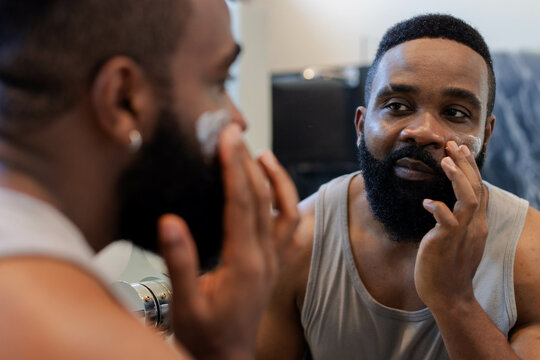 Applying facial cream African American man grooming beard at bathroom sink, with mirror and faucet
