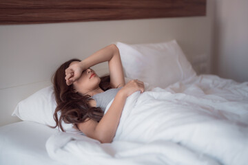 Obraz premium Young Asian woman lying in bed, covering her eyes with her arm, struggling with insomnia and attempting to find sleep during the early morning hours