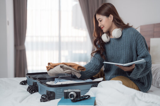 Young woman packing clothes in suitcase and checking list on notebook while sitting on bed at home, preparing for vacation trip or business trip