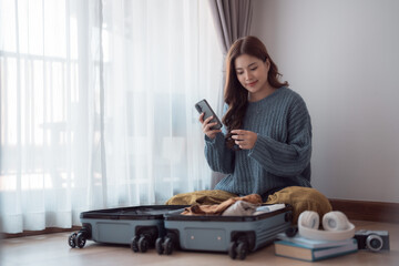 Young asian woman using mobile phone and packing clothes into suitcase, preparing for holiday...