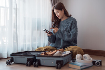 Young woman sitting beside a suitcase, checking her smartphone and smiling while packing clothes...