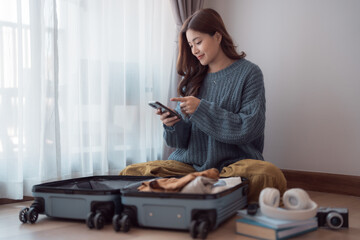 Happy asian woman packing clothes into travel bag, using mobile phone, preparing for vacation trip, sitting near window at home, booking hotel or tickets online