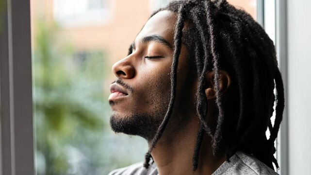 Young man with dreadlocks breathing deeply near window