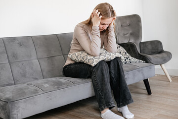 Young woman sitting on a sofa, holding her head with her hands, suffering from depression and...