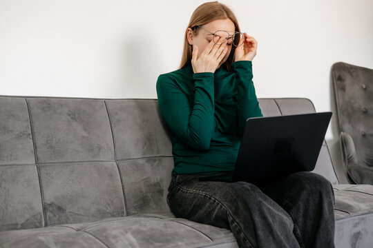 
Young woman sitting at desk, rubbing tired eyes from screen fatigue and stress. Concept of digital eye strain, overwork, computer vision syndrome, burnout, and remote work pressure 
