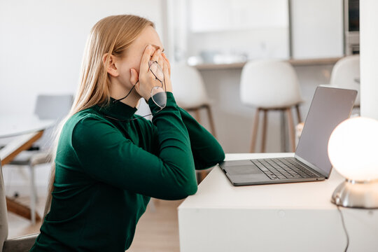 
Young woman sitting at desk, rubbing tired eyes from screen fatigue and stress. Concept of digital eye strain, overwork, computer vision syndrome, burnout, and remote work pressure 
