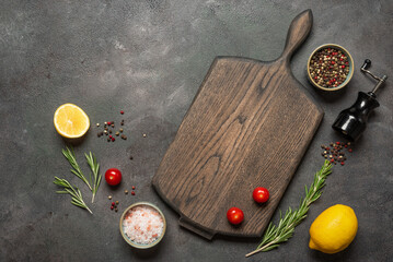 Culinary background with empty wooden cutting board, rosemary, peppercorns, pink salt, cherry tomatoes and lemon, dark rustic background, top view, flat lay