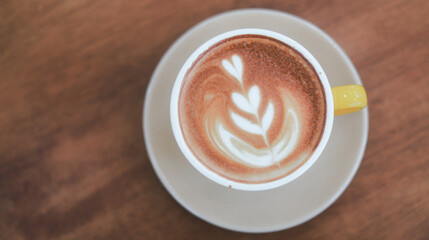 Close-up of hot cappuccino with beautiful latte art in yellow ceramic cup on wooden table.
